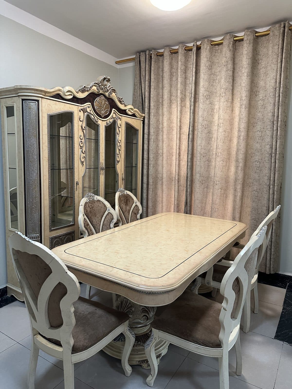 Dining table with carved cream chairs beside an ornate glass-fronted display cabinet and beige curtains
