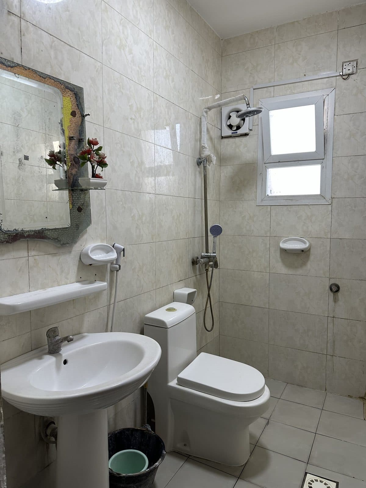 Cream-tiled bathroom with a basin, toilet, wall-mounted hand shower, a mirror with a small shelf and a window with an extractor fan