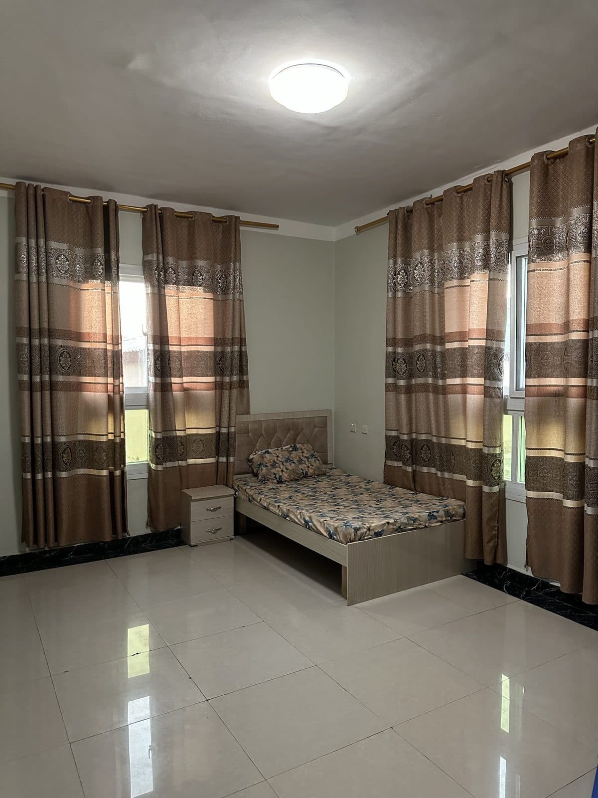 Second bedroom with a single bed, a tufted beige headboard, a small bedside cabinet and two curtained windows